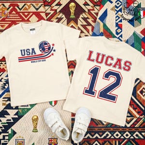 K&ouml;nnte beinhalten: Zwei cremefarbene T-Shirts mit Fu&szlig;ballmotiven. Eines zeigt "USA WORLD CUP 2026" mit einem Fu&szlig;ball-Design. Das andere hat den Namen "LUCAS" und die Nummer "12". Wei&szlig;e Turnschuhe sind unten zu sehen.