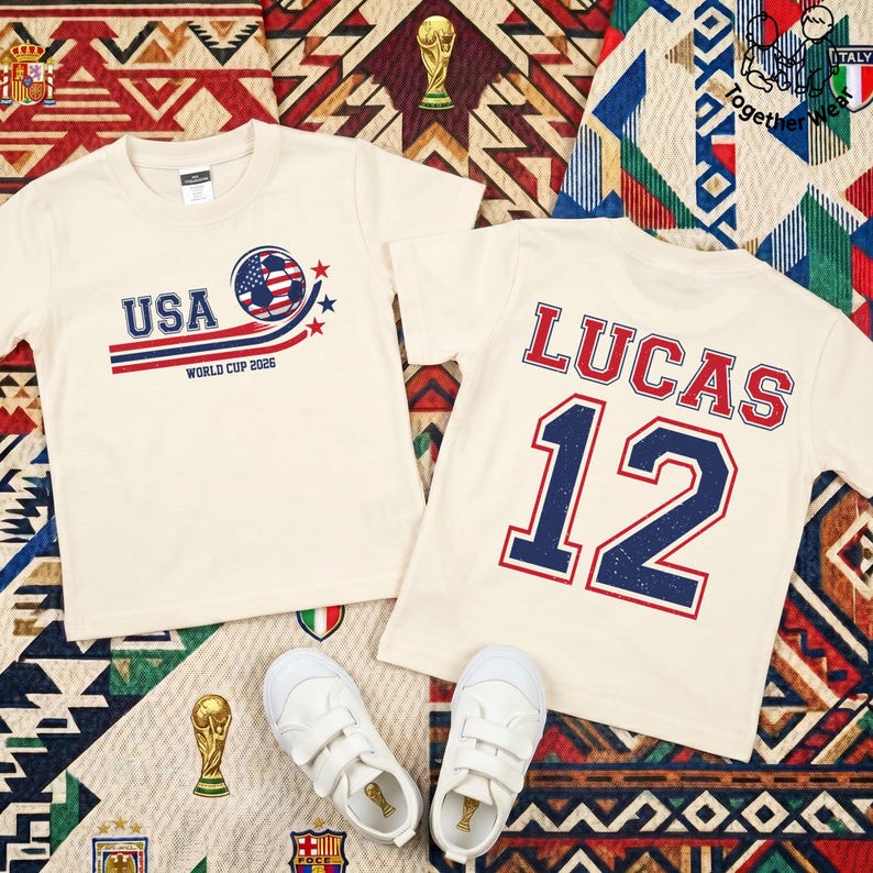 K&ouml;nnte beinhalten: Zwei cremefarbene T-Shirts mit Fu&szlig;ballmotiven. Eines zeigt "USA WORLD CUP 2026" mit einem Fu&szlig;ball-Design. Das andere hat den Namen "LUCAS" und die Nummer "12". Wei&szlig;e Turnschuhe sind unten zu sehen.