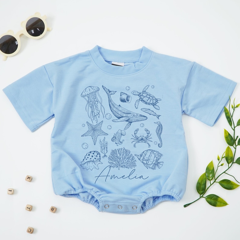 May include: Light blue baby romper with a sea life design, including a whale, jellyfish, and seahorse. The name "Amelia" is printed in a script font. The romper has short sleeves and snap closures.