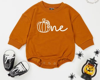 May include: An orange long-sleeved baby romper with a white pumpkin graphic and the word "one". The image includes black sneakers, a jack-o'-lantern, and plastic spiders, suggesting a Halloween theme.