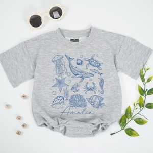 May include: A gray baby romper with short sleeves, featuring a blue illustration of various sea creatures, including a whale, jellyfish, and starfish. The name "Amelia" is written in cursive script below the illustration. Sunglasses are visible in the top left corner.