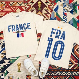 May include: Two cream-colored t-shirts featuring blue text and graphics. One shirt displays 'FRANCE SOCCER 2026' with a French flag icon. The other shirt shows 'FRA 10' with two orange stars above the text. A pair of white sneakers is placed below.