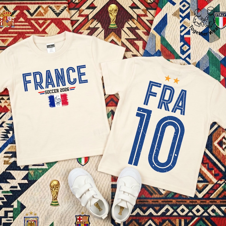 May include: Two cream-colored t-shirts featuring blue text and graphics. One shirt displays 'FRANCE SOCCER 2026' with a French flag icon. The other shirt shows 'FRA 10' with two orange stars above the text. A pair of white sneakers is placed below.