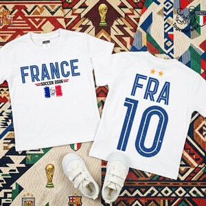 May include: Two white short-sleeved t-shirts. The left shirt features the blue text FRANCE, SOCCER 2026, and a French flag graphic. The right shirt displays the blue text FRA 10 with two orange stars above. A pair of white sneakers is placed below.
