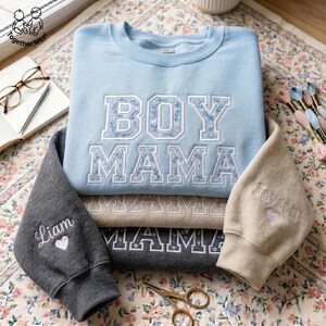 May include: A stack of embroidered sweatshirts in light blue, tan, and gray. The top sweatshirt says "BOY MAMA" in white and blue floral embroidery. The sleeves are embroidered with names and hearts. A pair of glasses, a notebook, and embroidery floss are in the background.