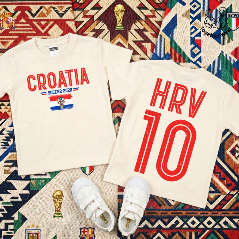 May include: Two cream-colored t-shirts featuring red text and graphics. One shirt displays CROATIA SOCCER 2026 with a Croatian flag. The other shirt features the text HRV and the number 10 in large red font. A pair of white sneakers is placed below.