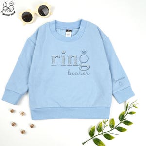 May include: A light blue sweatshirt with the words "ring bearer" embroidered in silver thread. The design includes a diamond ring graphic. The sleeve has the name "James" embroidered. Sunglasses and wooden letter blocks are in the background.