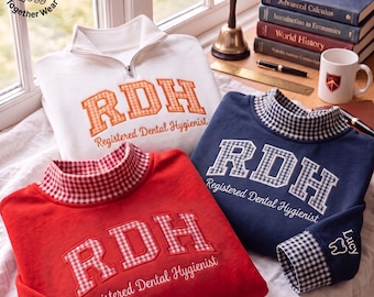 Puede incluir: Tres sudaderas en blanco, rojo y azul marino. Cada una presenta el texto bordado RDH Registered Dental Hygienist. La sudadera blanca tiene una cremallera de un cuarto, mientras que las rojas y azul marino tienen cuellos altos y puños de cuadros vichy.
