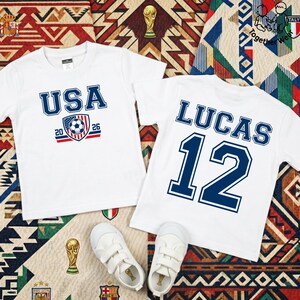 Może przedstawiać: Dwie białe koszulki z niebieskim tekstem i grafiką. Jedna koszulka m&oacute;wi "USA 2026" z grafiką piłki nożnej. Druga m&oacute;wi "LUCAS 12". Białe trampki poniżej. Koszulki leżą na wzorzystej powierzchni.