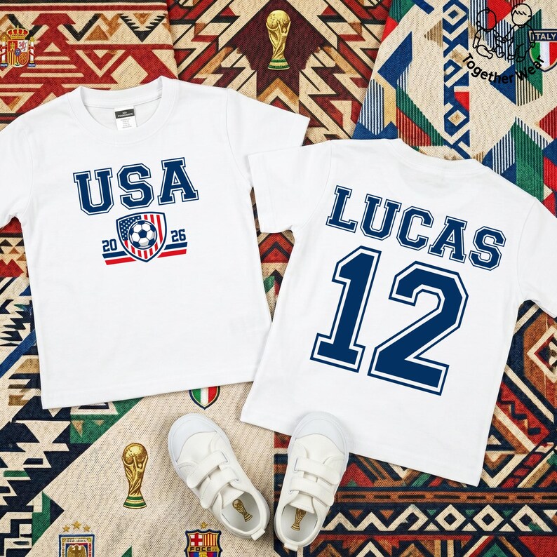 Może przedstawiać: Dwie białe koszulki z niebieskim tekstem i grafiką. Jedna koszulka m&oacute;wi "USA 2026" z grafiką piłki nożnej. Druga m&oacute;wi "LUCAS 12". Białe trampki poniżej. Koszulki leżą na wzorzystej powierzchni.
