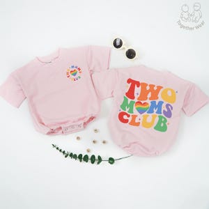 May include: Two pink baby rompers with short sleeves. One has a rainbow heart and "TWO MOMS CLUB" text on the front. The other has the same text on the back. Sunglasses are on the white surface.
