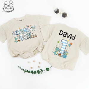 May include: Two beige baby rompers. One says "BIRTHDAY BOY" with colorful graphics. The other has "David" and a number "1" with a dinosaur, balloons, and a castle. Sunglasses and greenery are also in the image.