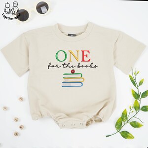 May include: A beige short-sleeved romper featuring the embroidered text ONE for the books in green, yellow, and red, with a stack of three books and a small red apple graphic centered below the text on the front of the garment.