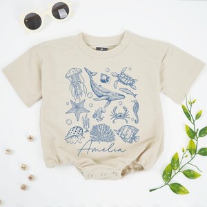 May include: A beige short-sleeved baby romper featuring a blue ocean-themed illustration with a whale, jellyfish, and other sea creatures. The name "Amelia" is written in cursive. Sunglasses are in the top left corner.