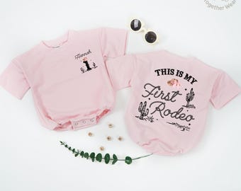 May include: Two pink baby bodysuits. One has the name "Hannah" with a graphic of a cowboy hat and cactus. The other says "THIS IS MY First Rodeo" with a cowboy hat, cactus, and rope design. Sunglasses are nearby.