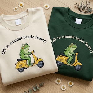 May include: Two crewneck sweatshirts, one cream and one dark green, each featuring an embroidered green frog riding a yellow scooter. The text "Off to commit bestie foolery" is arched above the frog on both garments, which have plain labels.