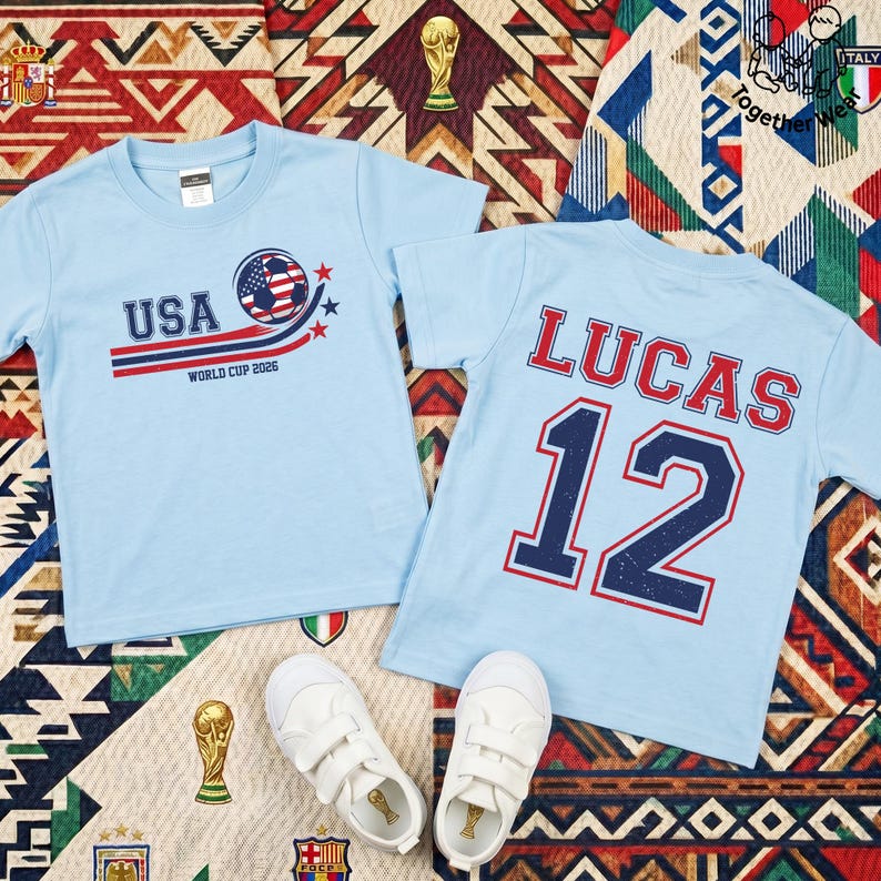 K&ouml;nnte beinhalten: Zwei hellblaue T-Shirts mit Fu&szlig;ballmotiven. Eines zeigt "USA" mit einem Fu&szlig;ball und "World Cup 2026". Das andere zeigt "LUCAS" und die Nummer "12". Wei&szlig;e Turnschuhe liegen unten auf einem gemusterten Hintergrund.