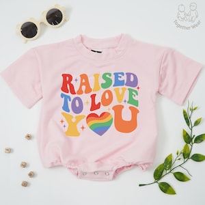 May include: A light pink baby romper with the text "RAISED TO LOVE YOU" in colorful, retro-style lettering. The design includes a rainbow heart and star accents. The romper has short sleeves and a round neckline.