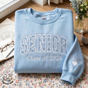 May include: A light blue sweatshirt with the word "SENIOR" embroidered across the chest in white floral lettering. The text "Class of 2026" is below. The sleeve has the name "Lauren" and a heart.