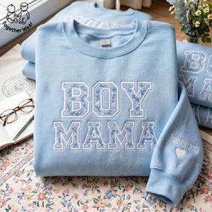 May include: Light blue sweatshirt with the embroidered text "BOY MAMA" in white floral lettering. The sleeve features the name "Jayson" and a heart. The sweatshirt is part of the "Together Wear" collection.
