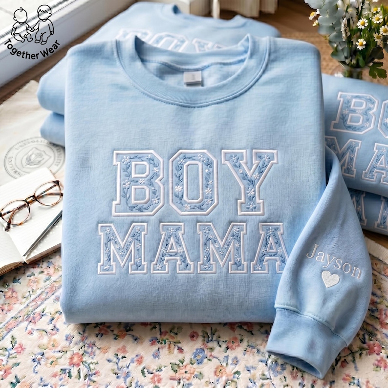 May include: Light blue sweatshirt with the embroidered text "BOY MAMA" in white floral lettering. The sleeve features the name "Jayson" and a heart. The sweatshirt is part of the "Together Wear" collection.