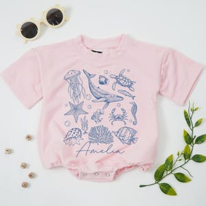 May include: A light pink baby romper with a crew neck and short sleeves. The romper is decorated with blue illustrations of sea creatures, including a whale, jellyfish, and starfish. The name "Amelia" is written in cursive script.