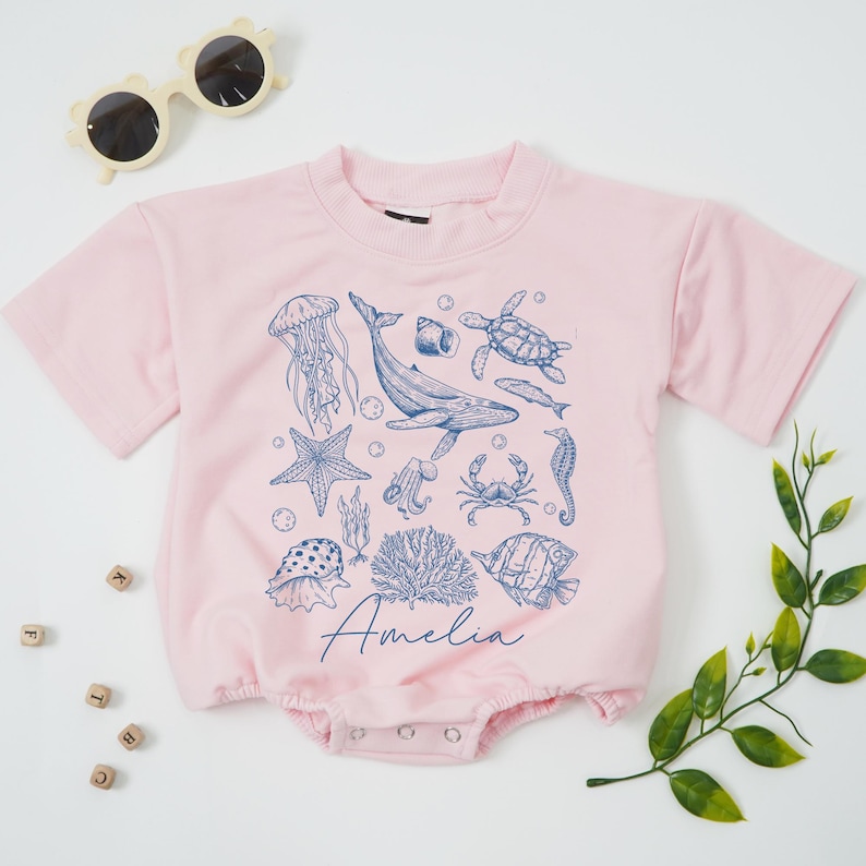 May include: A light pink baby romper with a crew neck and short sleeves. The romper is decorated with blue illustrations of sea creatures, including a whale, jellyfish, and starfish. The name "Amelia" is written in cursive script.