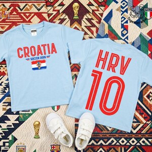 May include: Two light blue t-shirts. The left shirt features the red text "CROATIA" above "SOCCER 2026" and a Croatian flag graphic. The right shirt displays the red text "HRV" above the number "10". Two white sneakers are placed below the shirts.