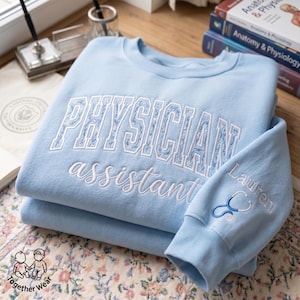 May include: A light blue sweatshirt with the words "PHYSICIAN assistant" embroidered in white. The sleeve features the name "Lauren" and a stethoscope design. The sweatshirt is folded on a floral patterned surface.
