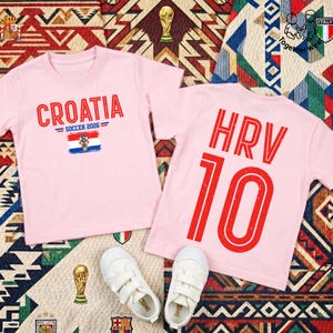 May include: Two light pink t-shirts. The left shirt features the text CROATIA SOCCER 2026 above a Croatian flag graphic. The right shirt displays the text HRV and the number 10 in large red font. A pair of white sneakers sits below the shirts.