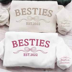 May include: Two sweatshirts, one cream and one white, with the word "BESTIES" embroidered across the front. The cream sweatshirt has brown embroidery, and the white sweatshirt has pink embroidery. Both have "EST.2022" and a bow design. The sleeves are embroidered with "Sophia Lauren".