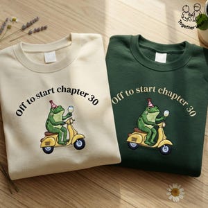 May include: A cream-colored sweatshirt and a dark green sweatshirt, each featuring a graphic of a green frog wearing a party hat, riding a yellow scooter, and holding a wine glass, with the text Off to start chapter 30 printed above.