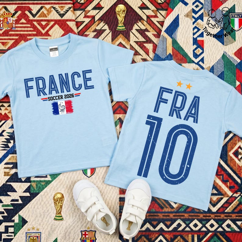 May include: Two light blue t-shirts. The left shirt features the text FRANCE SOCCER 2026 above a French flag graphic. The right shirt displays the text FRA 10 with two orange stars above it. A pair of white sneakers is placed below the shirts.