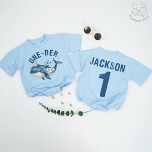 May include: Two light blue baby onesies. One features a whale graphic with the text "ONE-DER the SEA". The other has the name "JACKSON" and the number "1" printed on the back. Sunglasses and decorative elements are also present.