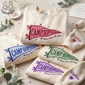 May include: Cream-colored sweatshirts with embroidered pennant designs. Each pennant reads "CAMP SOPHIA" with "Miami Beach, FL 2026" and a star. Sleeves are embroidered with "Sophia" and a heart. The sweatshirts are in red, blue, green, and purple.