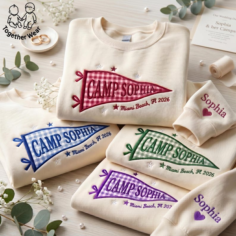 May include: Cream-colored sweatshirts with embroidered pennant designs. Each pennant reads "CAMP SOPHIA" with "Miami Beach, FL 2026" and a star. Sleeves are embroidered with "Sophia" and a heart. The sweatshirts are in red, blue, green, and purple.