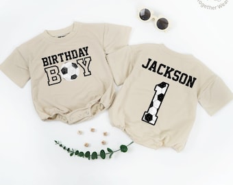 May include: Two beige baby bodysuits. One says "BIRTHDAY BOY" with a soccer ball graphic. The other says "JACKSON" with the number "1" in a soccer ball design. Sunglasses and decorative sprig are also present.