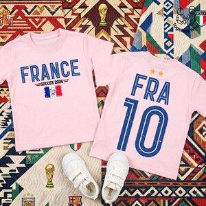 May include: Two light pink short-sleeved t-shirts. The left shirt features the text FRANCE SOCCER 2026 above a French flag graphic. The right shirt displays the text FRA 10 with two orange stars above it. Both shirts have blue distressed lettering.