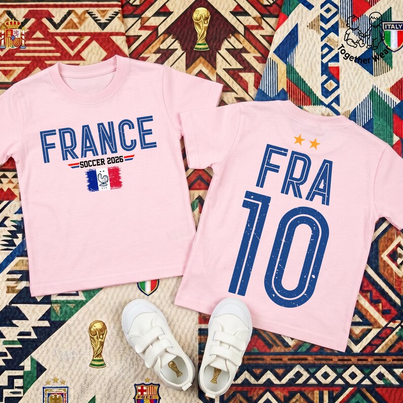 May include: Two light pink short-sleeved t-shirts. The left shirt features the text FRANCE SOCCER 2026 above a French flag graphic. The right shirt displays the text FRA 10 with two orange stars above it. Both shirts have blue distressed lettering.