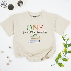 May include: A beige baby romper with the embroidered text "ONE for the books" in green, orange, and red. Below the text is a stack of books with a caterpillar on top. The romper has short sleeves and snap closures.
