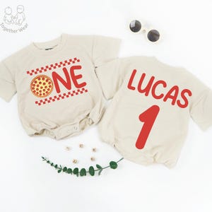 May include: Two beige baby rompers. One has the word "ONE" in red with a pizza graphic. The other has the name "LUCAS" and the number "1" in red. Sunglasses and decorative items are also present.