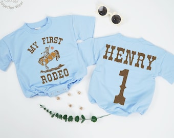 May include: Two light blue baby rompers. One says "MY FIRST RODEO" with a cowboy graphic. The other says "HENRY 1" in brown lettering. Sunglasses and decorative elements are also present.