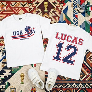 K&ouml;nnte beinhalten: Zwei wei&szlig;e T-Shirts mit Fu&szlig;ballmotiven. Eines zeigt "USA" mit einem Fu&szlig;ball-Grafik und "WORLD CUP 2026". Das andere zeigt "LUCAS" und die Nummer "12". Wei&szlig;e Turnschuhe sind ebenfalls im Bild auf einer gemusterten Oberfl&auml;che.