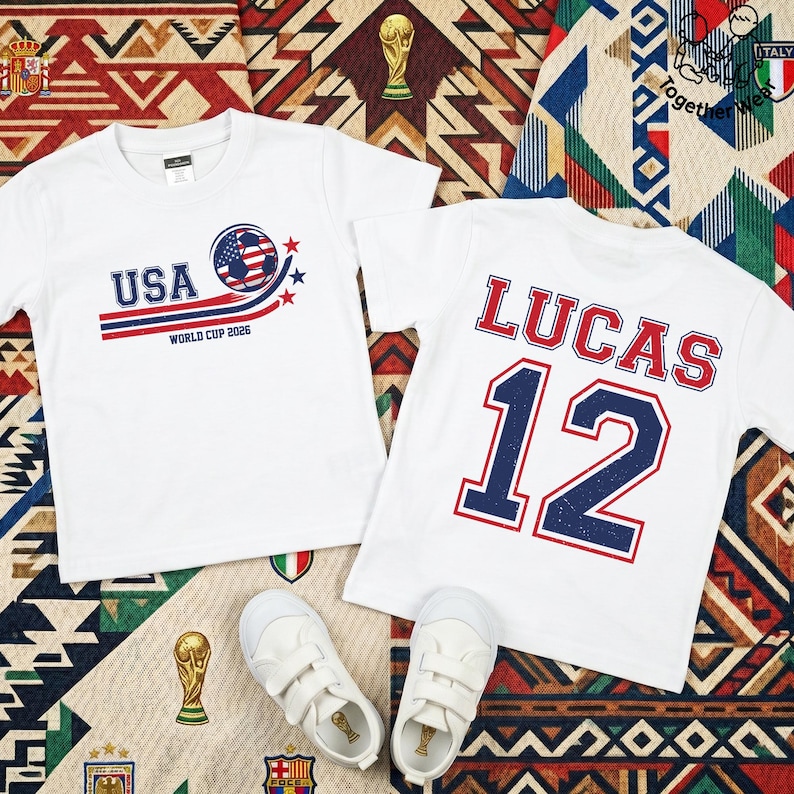K&ouml;nnte beinhalten: Zwei wei&szlig;e T-Shirts mit Fu&szlig;ballmotiven. Eines zeigt "USA" mit einem Fu&szlig;ball-Grafik und "WORLD CUP 2026". Das andere zeigt "LUCAS" und die Nummer "12". Wei&szlig;e Turnschuhe sind ebenfalls im Bild auf einer gemusterten Oberfl&auml;che.
