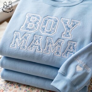 May include: A stack of light blue sweatshirts with the words "BOY MAMA" embroidered in white with floral detailing. The sleeve of one sweatshirt has the name "Jayson" and a heart embroidered in white. The brand logo is in the top left corner.