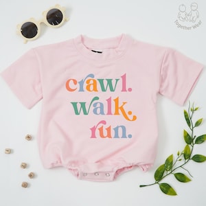 May include: A light pink baby romper with the words "crawl. walk. run." in colorful letters. The romper has short sleeves and a round neckline. A pair of cream-colored sunglasses and a sprig of greenery are also in the image.