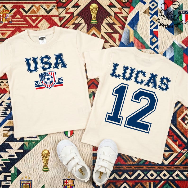 Może przedstawiać: Dwie kremowe koszulki z motywami piłkarskimi. Jedna z napisem "USA 2026" i grafiką piłki nożnej. Druga z imieniem "LUCAS" i numerem "12" w kolorze niebieskim. Białe trampki ułożone poniżej, na wzorzystej powierzchni.