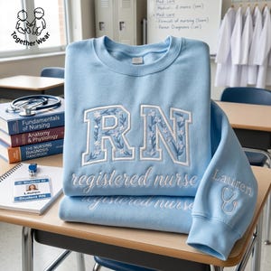 May include: A light blue sweatshirt with embroidered "RN" letters adorned with floral details. The words "registered nurse" are embroidered below. A stethoscope and the name "Lauren" are embroidered on the sleeve. Books, a student ID, and a stethoscope are also visible.