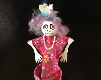 May include: A whimsical skeletal doll with purple hair, a pink floral dress, and a white pearl necklace. The doll is wearing a blue and white hat with flowers and is standing in a blue cone.