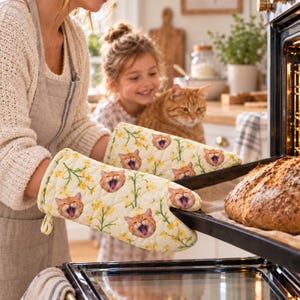 May include: A pair of quilted oven mitts featuring a repeating pattern of orange cat faces with open mouths and yellow floral sprigs on a cream background. The mitts are designed to protect hands while handling hot baking sheets from an oven.